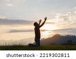 Young male kneeling down with hands open palm up praying to God on the mountain sunset background. 
