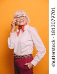 Small photo of Elderly smiling woman with gray hair and eyes wearing white shirt, orange cravat, red pants and leopard print belt, standing on isolated over orange background, talking on her phone