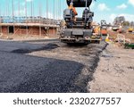 Small photo of Asphalt paver filled with hot tarmac laying new road surface on new residential housing development site and roadworker operator in orange hi-viz next to it