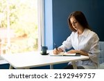 Serious female student writes a story in a notebook while sitting in a cafe with a cup of cappuccino. Focused young woman notes text in notepad