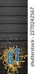 Small photo of Number 87 blue celebration candle and gold confetti on dark wooden background. 87th birthday card. Anniversary and birthday concept. Vertical banner. Copy space