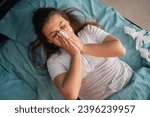 Small photo of Cold and flue concept. Sick woman suffering from running stuffy nose. Upset ill girl lying in bed, blowing her nose using paper napkin. Copy space. Top view
