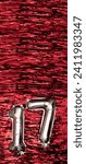 Small photo of Silver foil balloon number number 17 on a background of red tinsel decoration. Birthday greeting card, inscription seventeen. Anniversary event. Vertical Banner. copy space.