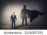 Small photo of Determined businessman stands confident in a hero stance and casting a brave superhero shadow on the wall behind. Business leadership and motivation concept. Ambition and strength symbols