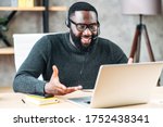 Smiling African-American guy uses a handsfree headset to talk online at his workplace, black confident man in glasses sits at the office desk and looks at laptop screen