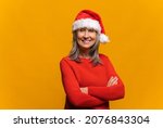 Happy confident senior grey-haired woman wearing red santa hat stands with arms crossed and looking at the camera isolated on yellow. Mature lady promoting new year deal, holiday sale