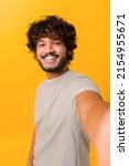 Small photo of Selfie time. Joyful carefree Indian young curly man talking selfie, looking at the camera and smiles, recording stories on his phone, vertical portrait