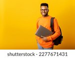 Small photo of Happy excited foreign male student with backpack standing isolated on yellow and holding laptop, arab guy in glasses ready to studying, back to school concept