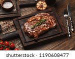 Grilled ribeye beef steak served on wooden board with rosemary, grilled garlic, fork and knife on wooden table