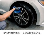 Car rims cleaning, car detailing wash concept. Cropped close up photo of male hand in black rubber glove with blue microfiber cloth washing car alloy wheel at car wash service