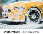 Cropped image of wheel of luxury yellow car in outdoors self-service car wash, covered with cleaning soap foam.