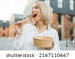 Happy positive business woman eating healthy salad on a break standing over city street background. Female dieting nutrition concept. Attractive smiling girl enjoying veggie meal.