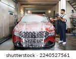 Side view portrait of young pleasant car wash worker, wearing protective overalls, cleaning automobile with high pressure water jet at car wash, spraying the cleaning foam. Car wash detailing.