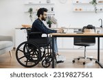 Small photo of Side view portrait of indian male in wheelchair working at writing desk in open-plan kitchen of modern apartment. Young adult with disability starting working day with checking email account at home.