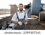 Serious construction worker in overalls and gloves standing and using modern tablet outdoors. Caucasian professional man surfing internet while working with devices on roof of factory.