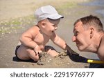 A small tanned boy in a white cap sits on the shore of a sandy beach and smiles at his dad. Close-up portrait. Dad and baby on the sea, family vacation concept