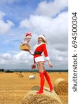 Small photo of A young farmhand girl dresses up in a Santa Claus suit and stands in front of a hay bale in the farmer's field. She decorates the hay bale with Christmas presents and smiles at the camera.