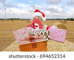 Small photo of A young farmhand girl dresses up in a Santa Clause suit and stands in front of a hay bale in the farmer's field. She decorates the hay bale with Christmas presents and smiles behind them.