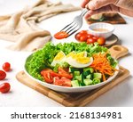Hand Holding a Fork with Cherry Tomato. Healthy Salad of Fresh Green Coral Lettuce, Cherry Tomato, Cucumber, Carrot and Egg on a Cutter Board and White background
