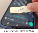 Small photo of Singapore - Oct 2023: A person placing a note showing the word SCAM on a mobile phone with potential scam message shown on mobile phone