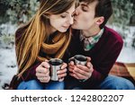 Portrait of happy young couple at the picnic on the Valentines Day in a snowy park. Man kiss girl, hold cups of mulled wine, hot tea, coffee. Christmas holiday, celebration. Happy new year.
