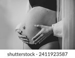 Small photo of Closeup belly of a woman. Pregnancy motherhood procreation concept. Pregnant female waiting for newborn baby. Young girl touching and holding belly, caring about health indoors. Black and white photo