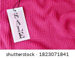 Word Sale on price tag on pink knitted wool fabric with folds or sweater. A gray textured cardboard label card attached to the black tape on the clothes. Shopping concept, background with copy space.