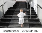 Back view of cute funny caucasian blond kid boy wearing white pool spa bathrobe walking upstairs on big marble staircase. Alone small child meet challenge and purpose. Rich luxury lifestyle concept