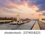 Small photo of Scenic view od big cargo freight tugboat barge boat ship sailing by german inland canal Magdeburg bridge against warm sunset sky background. Water transportation logistics shipping industry Europe