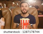 Excited male viewer keeping popcorn in hands and watching interesting film in cinema. Young bearded man wearing shirt expecting final of movie. Concept of entertainment and leisure.