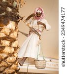 Small photo of Old Saudi farmer harnessed on top of a palm tree to pick up dates and lower them down in the basket,05-08-2023,Qassim,Saudi Arabia