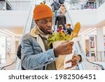Small photo of African man with a paper bag of groceries looks surprised and upset at a receipt from a supermarket with high prices against the background of an escalator with customers in the shopping center. The