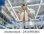 Man in store chooses drink. Guy in supermarket reads label, composition of sodas, chooses, compares prices, looks for benefits. Bottom view from grocery cart. Racks with huge variety of drinking goods