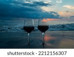 Close-up of two glasses of red wine standing in the sand against the background of the waves of the sea with a beautiful sunset. Beautiful picture of a romantic summer evening at the sea