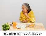 Small photo of Senior asian woman with acid reflux problem, sore throat. While eating patty, food stuck in the throat, burning sensation in the esophagus, irritation, coughing, suffocation and difficulty swallowing.
