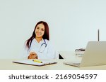 Small photo of Portrait kind hearted Asian female doctor specializing in medical field sits and works in the hospital office smiling at the camera with goodwill.