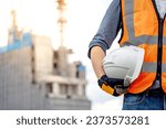 Small photo of Safety workwear concept. Male hand holding white safety helmet or hard hat. Construction worker man with reflective orange vest and protective gloves standing at unfinished building with tower crane