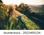 Young woman traveler with long loose curly hair sits on green grass meadow with flowers and types on smartphone against hilly landscape under sunlight
