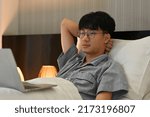 Small photo of Portrait of Asian male in nightdress watching an online streaming movie on computer laptop before he sleeps.
