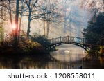 Scenic view of misty autumn landscape with beautiful old bridge with swan on pond in the garden with red maple foliage.
