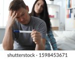 Small photo of Portrait of anxious worried man holding pregnancy test in hand. Troublous female sitting behind and supporting male. People in cozy bedroom. Unexpected pregnancy concept