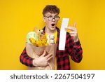 young shocked shopper with a package of groceries holding a check and being surprised by the high prices on a yellow background, the concept of a rise in price