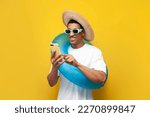 Small photo of cheerful young guy afro american with inflatable swim ring uses smartphone on yellow isolated background, man in summer on beach is typing on phone, concept of travel and summer holidays