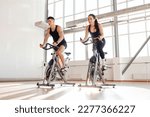 Small photo of beautiful athletic couple of cyclists train in the fitness room on static bicycle simulator, man trainer and fitness woman go in for sports together on simulators in the morning in bright room