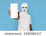 Small photo of african american man thug in white balaclava shows empty smartphone screen on blue isolated background, hoodlum in mask advertises copy space on phone