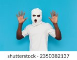 Small photo of I give up. shocked african american male thug in white balaclava raising hands on blue isolated background, caught bully in mask in amazement with open mouth showing palms