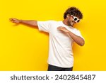 Small photo of young indian guy in sunglasses dancing dab on yellow isolated background, south asian man in white t-shirt raises his hands up and shows dab gesture