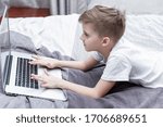 A boy of 8 years old is lying on the bed and uses a laptop. Distance learning during the coronavirus pandemic. 