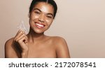 Small photo of Smiling african american woman holds gua sha scrapper tool for facial lifting, tcm face cosmetic treatment, home spa procedure, standing over brown background