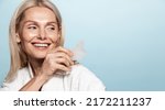 Small photo of Elegant smiling woman, 50 years old, using guasha tcm massaging tool, holding scrapper and looking aside at copy space, blue background